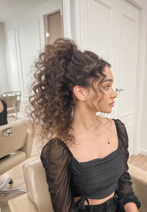 A woman with curly hair sitting in a chair, wearing a ponytail em cabelo cacheado hairstyle.