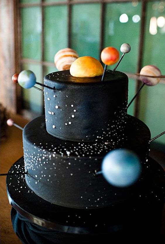 A Quinceanera-themed layer cake with a black base and planets on top, surrounded by cupcakes.