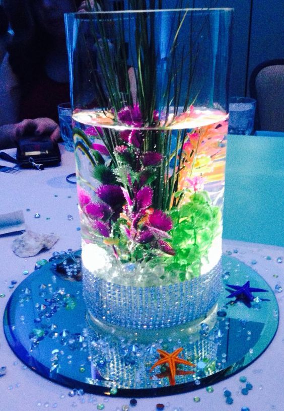 A Quinceanera centrepiece featuring a vase filled with water and flowers on top of a table, resembling a Little Mermaid theme.