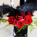 A Quinceanera table centerpiece featuring a vase filled with red roses and black feathers