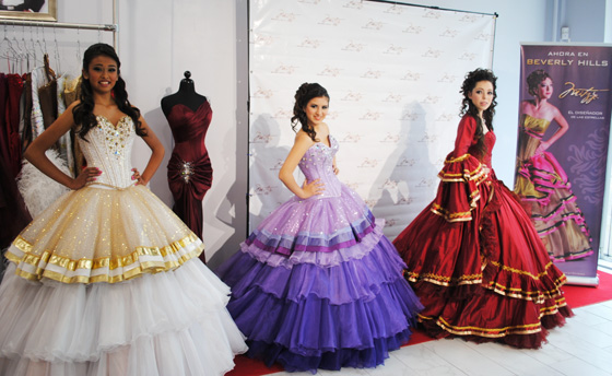 A group of women standing next to each other in Quinceanera dresses, posing for a photo with a mariachi band playing in the background.