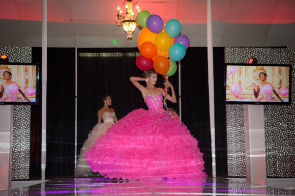 A woman in a pink Quinceanera gown holding a bunch of Quinceanera balloons in a function hall.