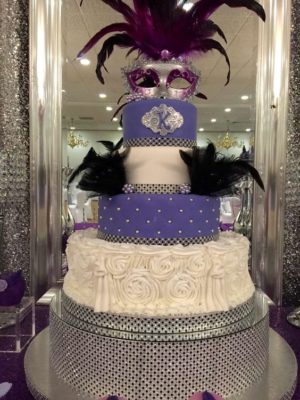 An image of a masquerade themed Quinceanera. The Quinceañera dresses are beautifully displayed. In the background, there is a three-tiered cake adorned with purple and black decorations.