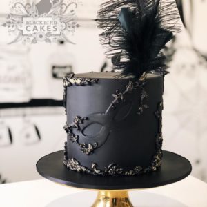 A Quinceanera cake decorated with black and gold colors and topped with a feather.