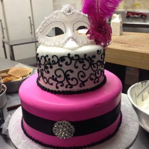 A Quinceanera cake decorated with pink and black colors. The cake has a mask on top.
