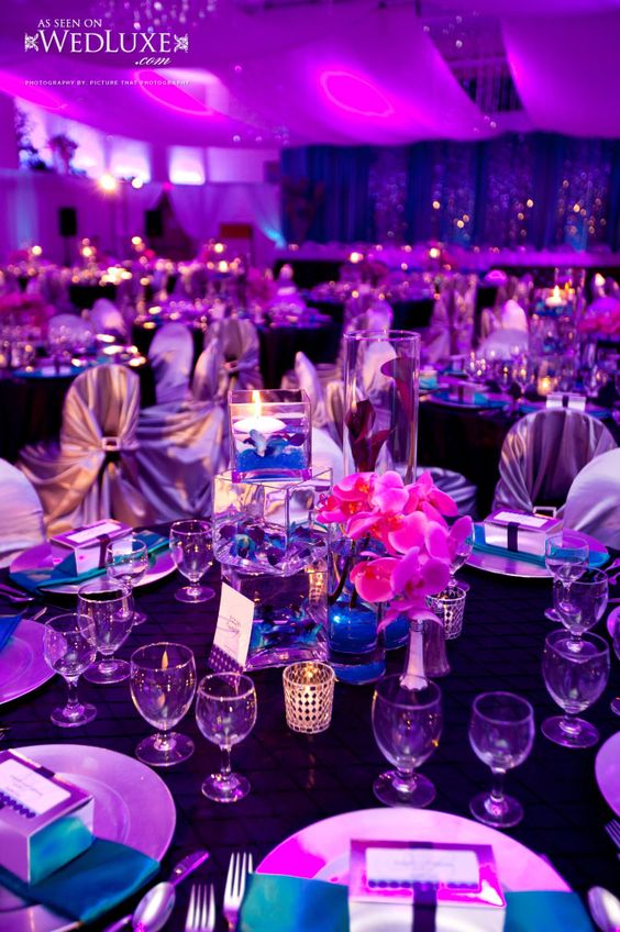 A table set for a Quinceanera reception with royal blue, gold, and purple lighting, featuring a Quinceanera invitation.