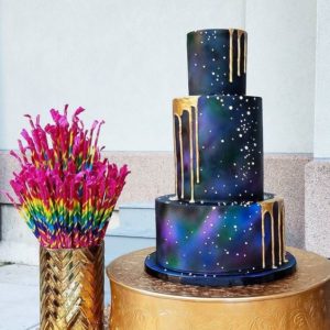 A Quinceanera themed cake, with a gold galaxy design, consisting of three tiers and placed on a table.