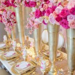 A Quinceanera-themed image featuring floral design. The image shows a long table topped with gold vases filled with pink flowers.