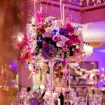 Quinceanera image of a tall vase with flowers and candles on a table, with a floral design in a function hall.