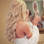 Curly hair with pieces pulled back, a woman in a white dress looking at her reflection in a mirror at a Quinceanera event