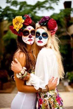 Two women in Quinceanera themed Day of the Dead costumes hugging each other, portraying dia de los muertos make up.