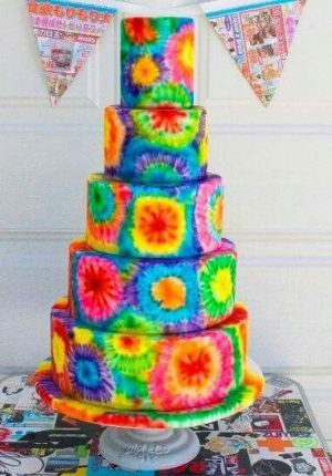 A colorful Quinceanera cake with rainbow tie dye design, adorned with purple flowers on top