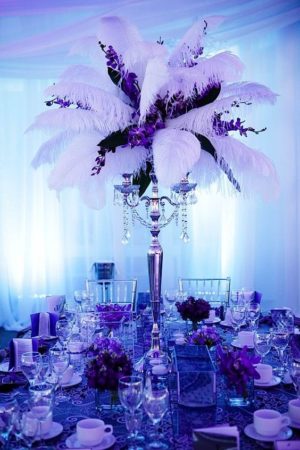 A beautiful quinceanera table setting with a blue and purple theme. The centerpiece is tall and features a combination of blue and purple flowers. The table is set with purple and white tableware, creating an elegant and vibrant atmosphere.