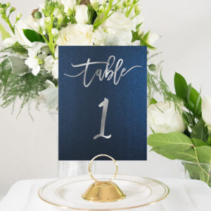 Gold and black tables with a blue table number featuring a gold bell on a white table for a Quinceañera.