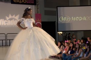 A woman in a white dress walking on a runway at a Quinceanera fashion show