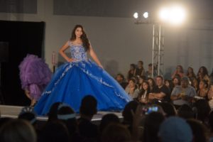 Quinceanera fashion show featuring haute couture. A woman in a blue dress is walking down the runway.