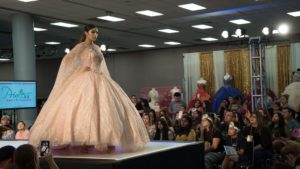 A woman in a Quinceanera gown walking down a runway during a fashion show.