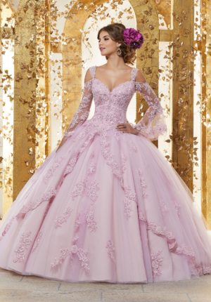 A woman in a pink ball gown with bell sleeves standing in front of a gold wall, showcasing Quinceañera dresses.