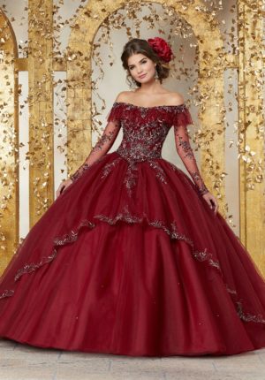 A woman in a red ball gown standing in front of a gold wall, wearing dress 89235 Vizcaya by Morilee for Quinceanera