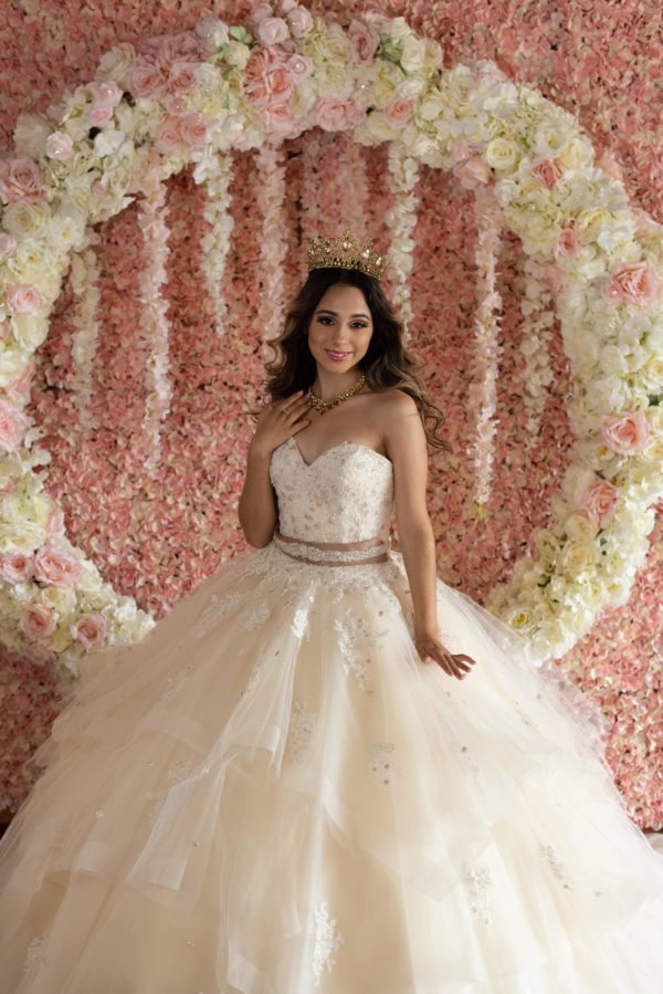 A woman in a Quinceanera dress standing in front of a floral wall, surrounded by Quinceanera decorations