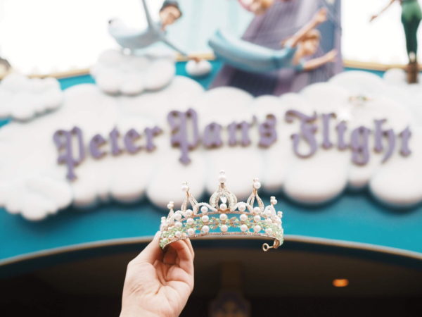 Girl's hand holding up a TInkerbell tiara with the Peter Pan ride in the background