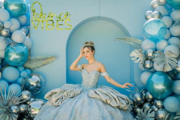 A woman in a ball gown standing in front of blue Quinceañera balloons and Quinceañera dresses