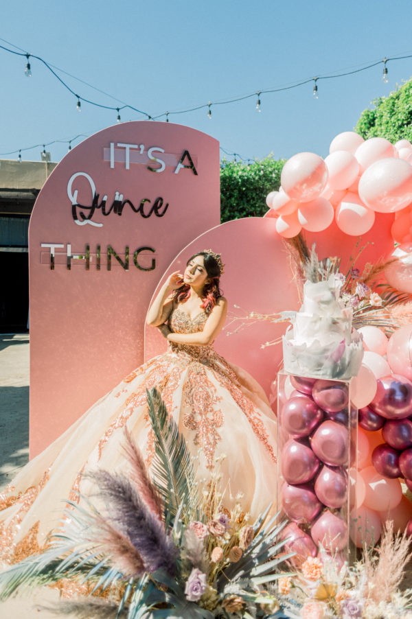 A woman in a Quinceanera ball gown standing next to a sign, surrounded by flower Quinceañera dresses