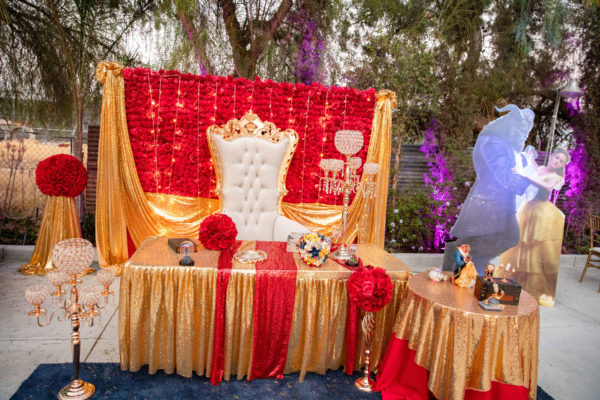 Quinceanera celebration in a function hall with a decorated stage featuring a gold chair and red flowers