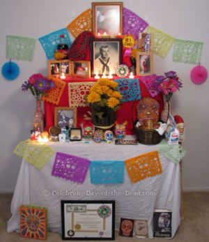 A Quinceanera-themed image featuring a DIY Day of the Dead altar. The altar is a table decorated with a tablecloth adorned with flowers.