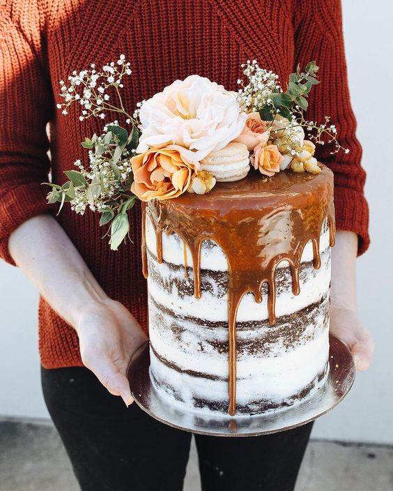 A Quinceanera cake with caramel dripping and a naked design. A woman is holding the cake, which has beautiful flowers on top.