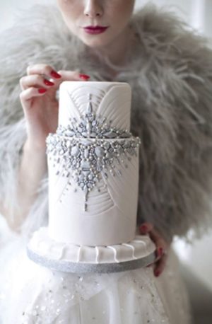 A Quinceanera cake, featuring bling and a winter theme. The cake is held by a woman wearing a white dress.