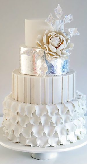 A three tiered white Quinceanera cake with a flower on top, adorned with silver decor