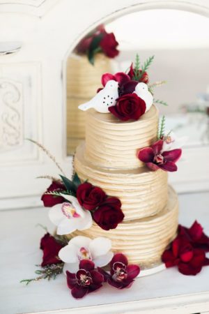 A gold Quinceanera cake with red flowers on top, featuring a burgundy and blue color palette