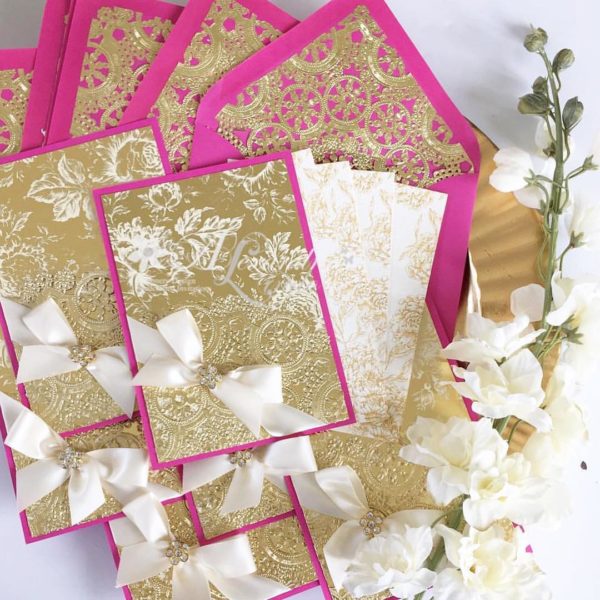 A Quinceanera-themed image featuring a petal textile background with a bunch of pink and gold cards adorned with white bows.