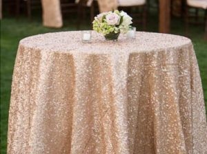 A Quinceanera-themed table decorated with gold sequins table covers and a vase of flowers on top