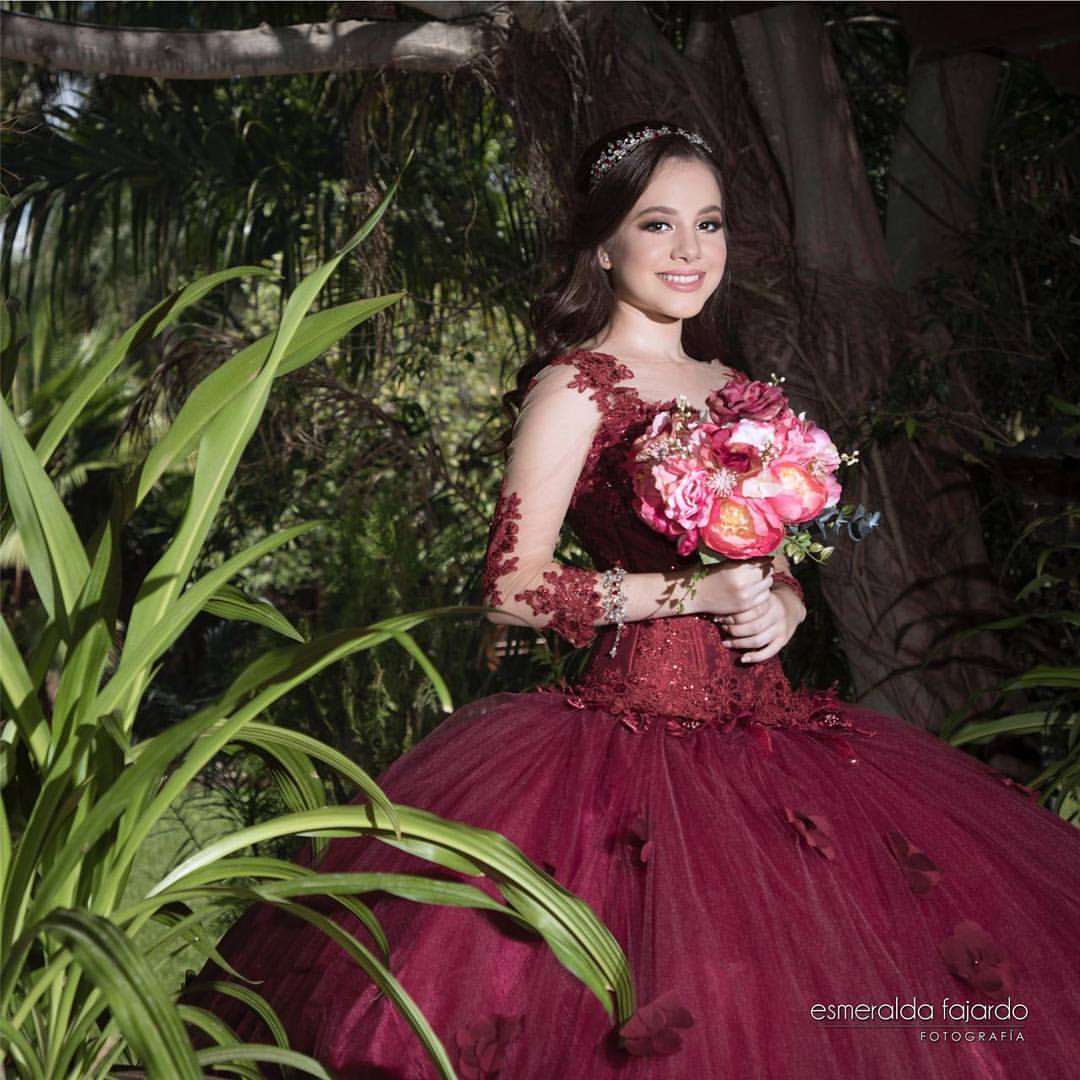 A woman in a red Quinceañera dress, holding a bouquet of flowers, with long sleeves, in the color burgundy