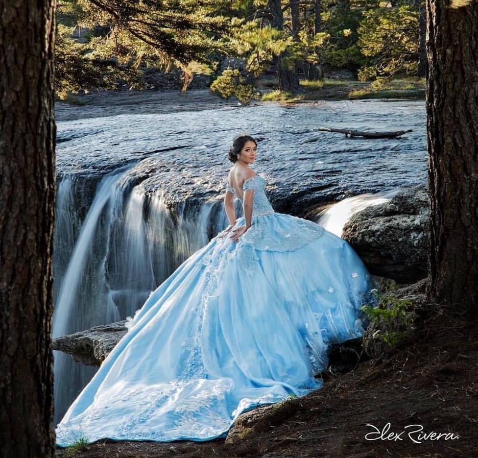 A woman in a blue dress sitting in front of a waterfall wearing one of the most elegant Quinceañera dresses